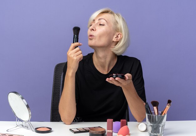 Pleased young beautiful girl sits at table with makeup tools applying powder blush with powder brush isolated on blue wall