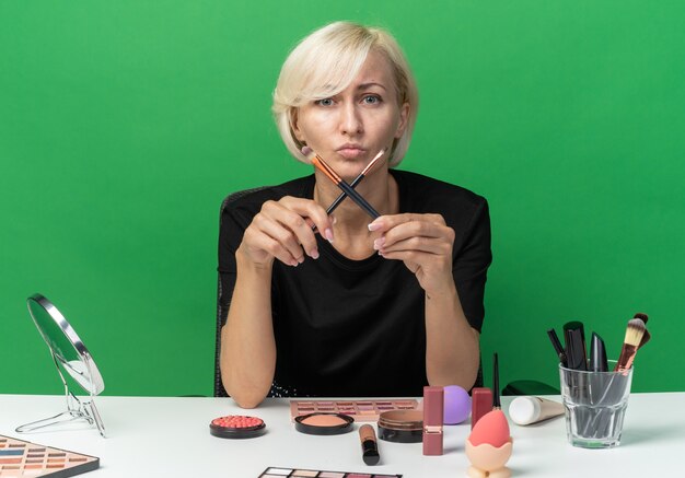 Pleased young beautiful girl sits at table with makeup tools holding and crossing makeup brushes isolated on green wall