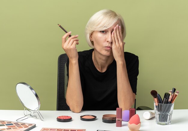 Pleased young beautiful girl sits at table with makeup tools holding eyeliner covered eye with hand isolated on olive green wall