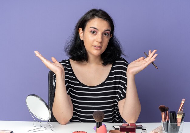 Pleased young beautiful girl sits at table with makeup tools holding eyeliner spreading hands isolated on blue wall