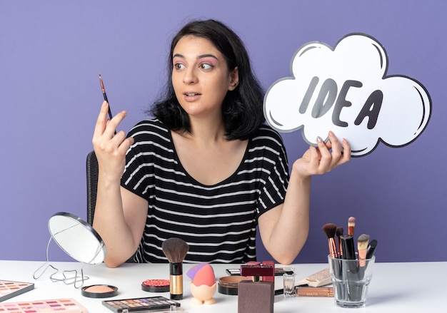 Foto gratuita la giovane bella ragazza piacevole si siede alla tavola con gli strumenti di trucco che tengono la bolla di idea con la spazzola di trucco isolata sulla parete blu