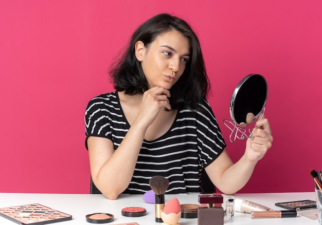 Pleased young beautiful girl sits at table with makeup tools holding and looking at mirror putting hand under chin isolated on pink wall