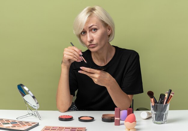 Pleased young beautiful girl sits at table with makeup tools holding makeup brush isolated on olive green wall