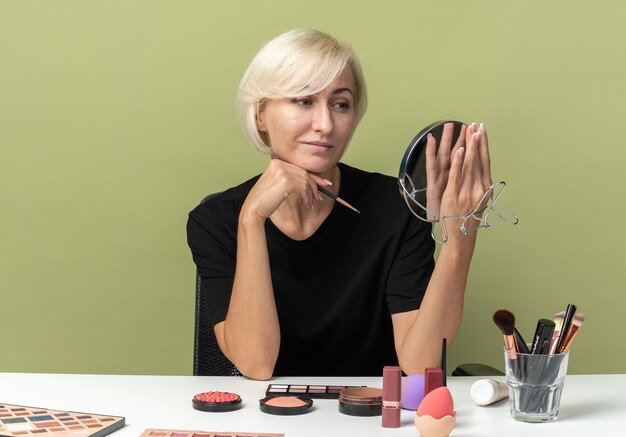 Pleased young beautiful girl sits at table with makeup tools holding makeup brush with mirror isolated on olive green wall