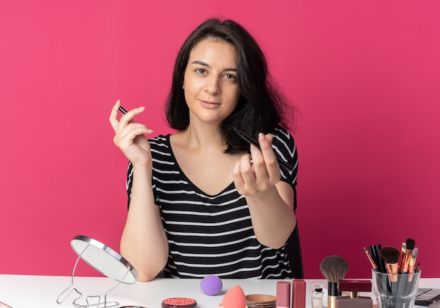 Pleased young beautiful girl sits at table with makeup tools holding out eyeliner  isolated on pink wall