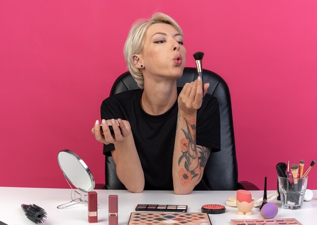 Pleased young beautiful girl sits at table with makeup tools holding powder blush with powder brush isolated on pink wall