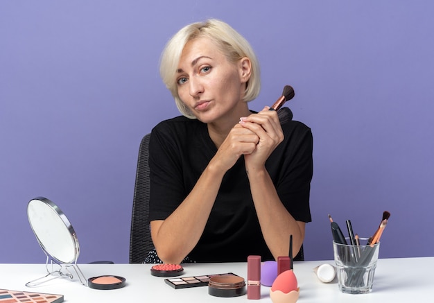 Pleased young beautiful girl sits at table with makeup tools holding powder brushes isolated on blue wall