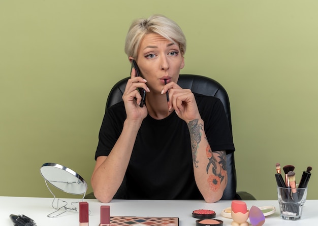 Pleased young beautiful girl sits at table with makeup tools speaks on phone holding makeup brush isolated on olive green wall