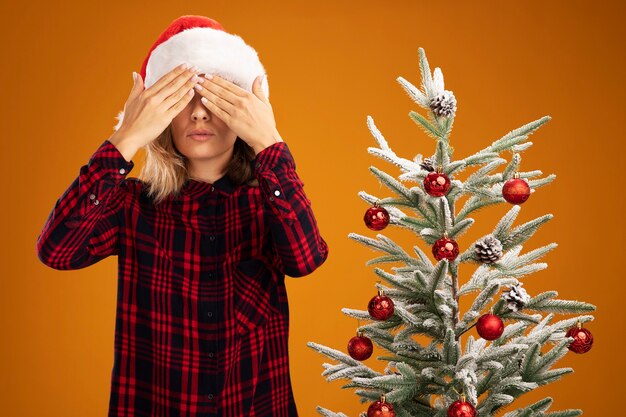 Pleased young beautiful girl standing nearby christmas tree wearing christmas hat covered eyes with hands isolated on orange background
