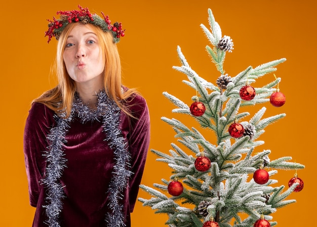 Lieta giovane bella ragazza in piedi vicino all'albero di natale che indossa un abito rosso e corona con ghirlanda sul collo che mostra gesto di bacio isolato su sfondo arancione