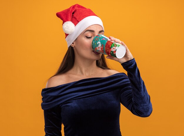 Pleased young beautiful girl wearing blue dress and christmas hat drinks from christmas cup isolated on orange background