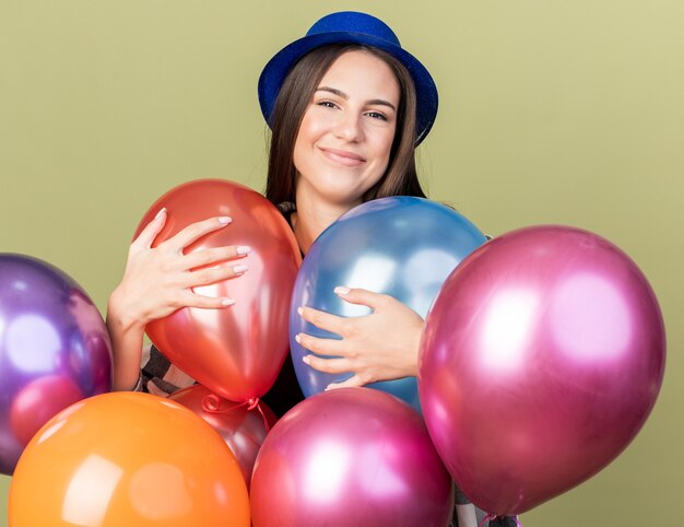 Pleased young beautiful girl wearing blue hat standing behind balloons 