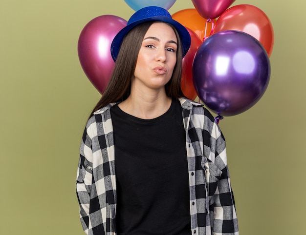 Free photo pleased young beautiful girl wearing blue hat standing in front balloons