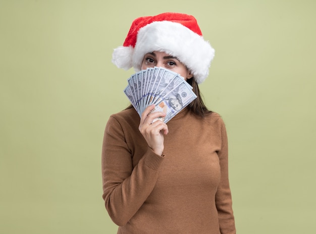 Pleased young beautiful girl wearing christmas hat covered face with cash isolated on olive green background