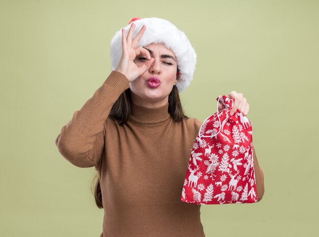 Pleased young beautiful girl wearing christmas hat holding christmas bag showing look gesture isolated on olive green background