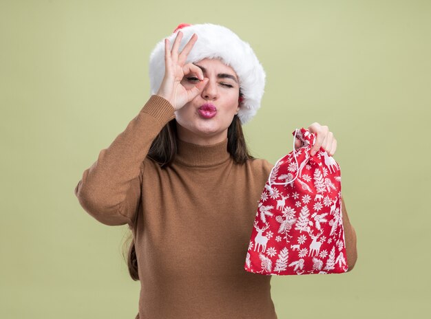 オリーブグリーンの背景に分離された外観のジェスチャーを示すクリスマスバッグを保持しているクリスマス帽子をかぶって喜んで若い美しい少女
