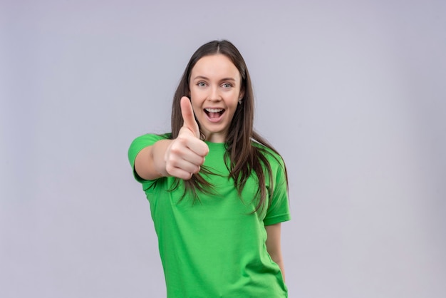 Довольная молодая красивая девушка в зеленой футболке, весело улыбаясь, показывает палец вверх, стоя на изолированном белом фоне