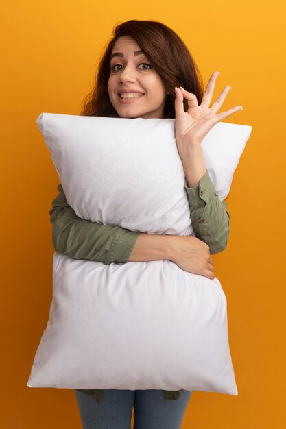 Pleased young beautiful girl wearing olive green t-shirt hugged pillow showing okay gesture isolated on yellow wall