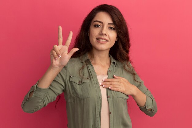 Pleased young beautiful girl wearing olive green t-shirt showing peace gesture putting hand on heart isolated on pink wall