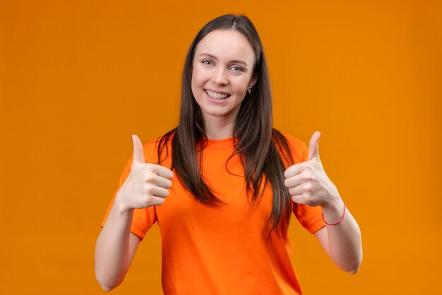 Pleased young beautiful girl wearing orange t-shirt smiling cheerfully showing thumbs up with both hands standing over isolated orange background