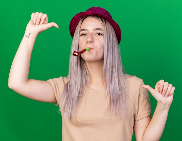 Pleased young beautiful girl wearing party hat blowing party whistle points at herself 