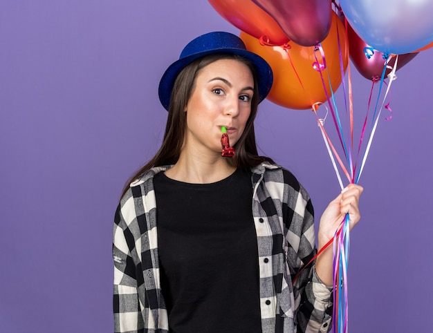 Довольная молодая красивая девушка в шляпе для вечеринки, держащая воздушные шары, дует свисток для вечеринки