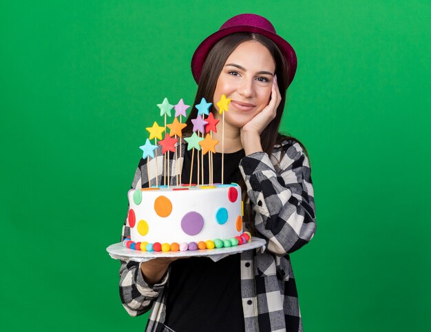 Pleased young beautiful girl wearing party hat holding cake putting hand on cheek 