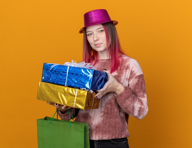 Pleased young beautiful girl wearing party hat holding gift bag with gift boxes isolated on orange wall
