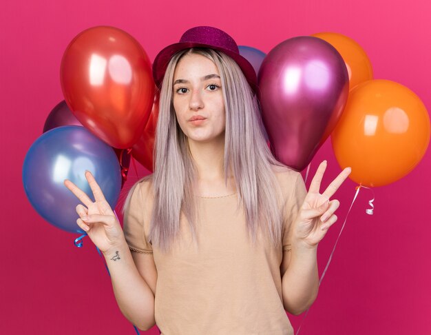 Pleased young beautiful girl wearing party hat standing in front balloons showing peace gesture 