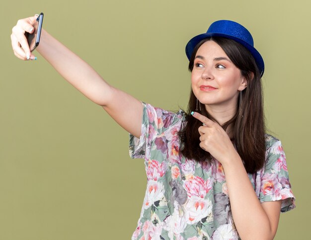 Довольная молодая красивая девушка в шляпе делает селфи на телефоне, изолированном на оливково-зеленой стене