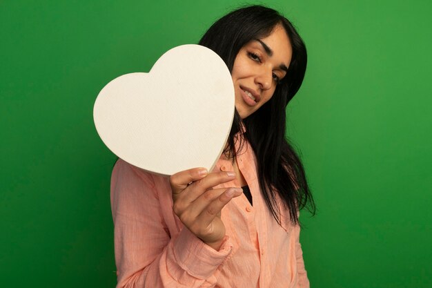 Pleased young beautiful girl wearing pink t-shirt holding heart shape box isolated on green