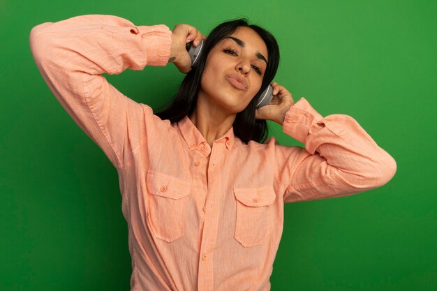 Довольная молодая красивая девушка в розовой футболке с наушниками изолирована на зеленой стене