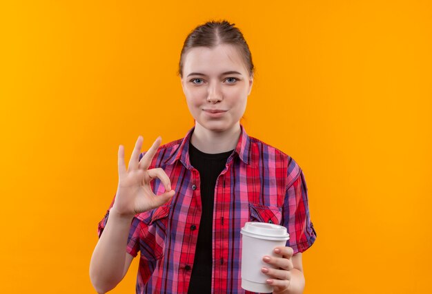 Довольная молодая красивая девушка в красной рубашке, держащая чашку кофе, показывая жест окей на изолированном желтом фоне с копией пространства