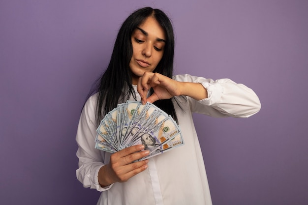 Free photo pleased young beautiful girl wearing white t-shirt holding and looking at money isolated on purple