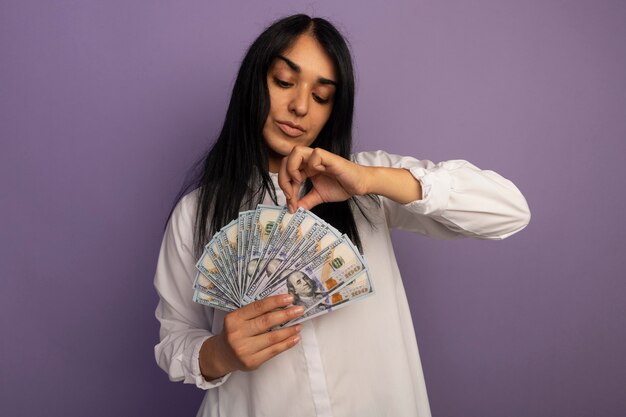 Pleased young beautiful girl wearing white t-shirt holding and looking at money isolated on purple