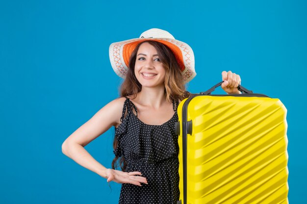 Pleased young beautiful traveler girl in dress in polka dot in summer hat holding suitcase looking at camera smiling cheerfully happy and positive standing over blue background