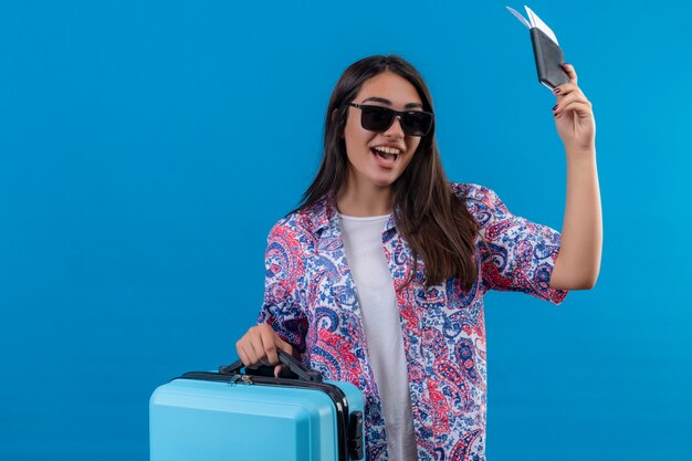 Pleased young beautiful traveler woman wearing sunglasses holding blue suitcase and tickets smiling cheerfully with happy face standing over blue background