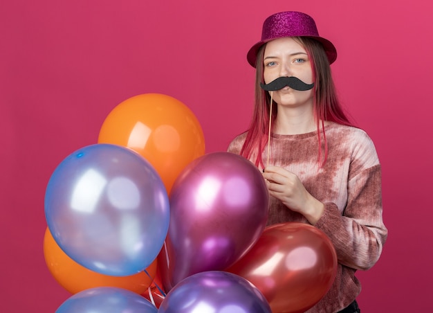 Free photo pleased young beautiful wearing party hat holding balloons with fake mustache on stick