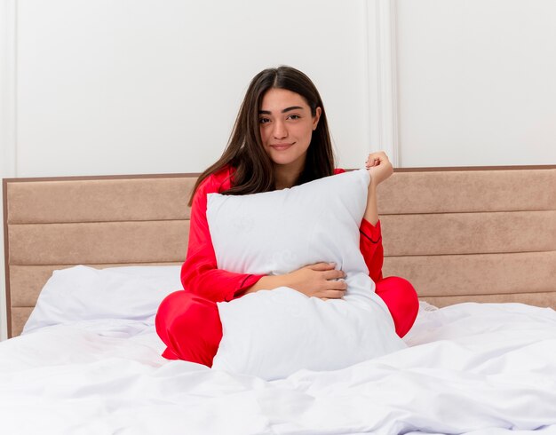 Pleased young beautiful woman in red pajamas sitting in bed with pillow smiling with happy face in bedroom interior 