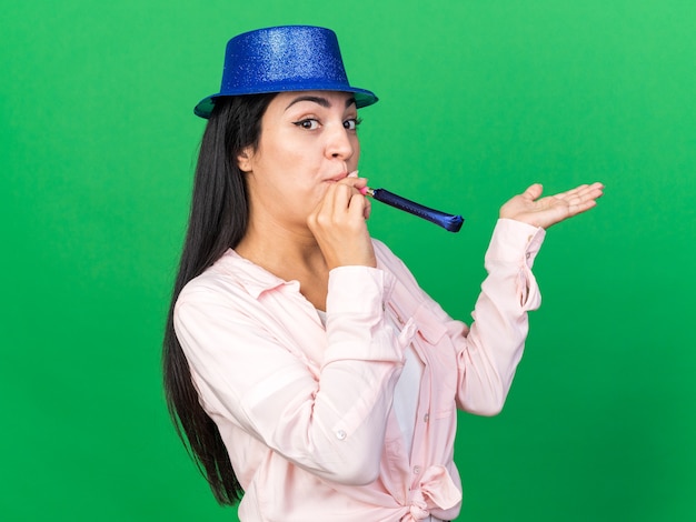 Lieta giovane bella donna che indossa cappello da festa che soffia fischio di festa spargendo la mano isolata sul muro verde