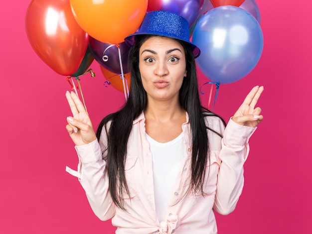 Pleased young beautiful woman wearing party hat standing in front balloons points at differents sides isolated on pink wall