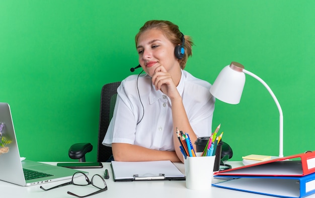 Pleased young blonde call centre girl wearing headset sitting at desk with work tools keeping hand on chin looking at laptop isolated on green wall