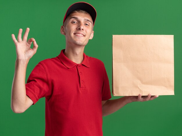 Довольный молодой блондин посыльный жестикулирует знак рукой и держит бумажный пакет