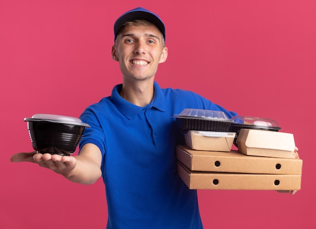 복사 공간이 있는 분홍색 벽에 격리된 피자 상자에 음식 용기와 패키지를 들고 있는 행복한 금발 배달 소년