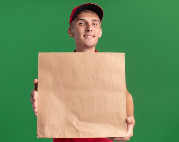 Pleased young blonde delivery boy holding paper package on green 