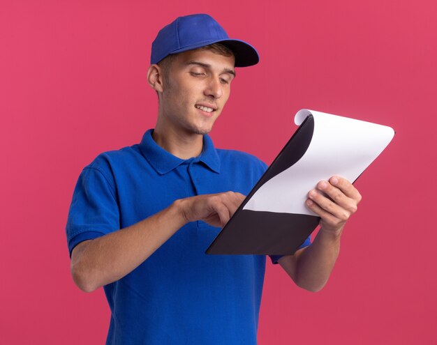 Pleased young blonde delivery boy holds and looks at clipboard on pink