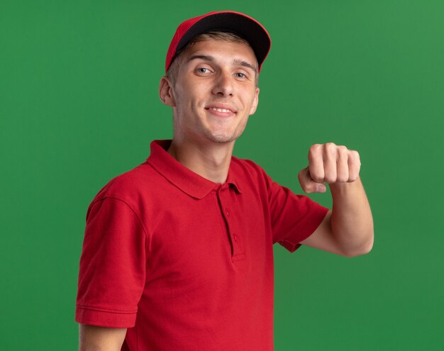 Foto gratuita il giovane ragazzo di consegna biondo piacevole tiene il pugno ed è isolato sulla parete verde con lo spazio della copia