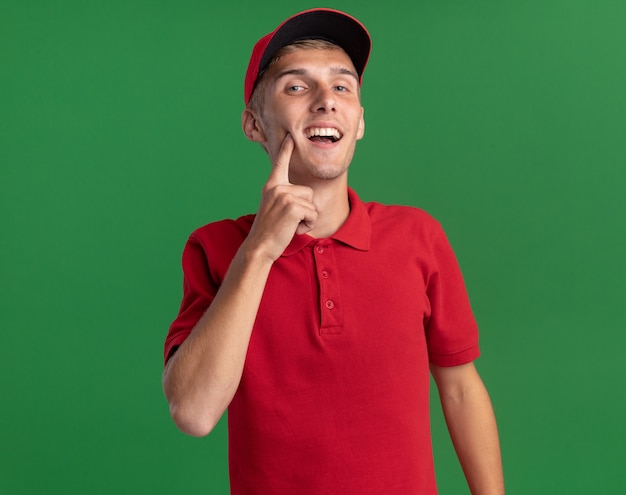 Pleased young blonde delivery boy puts finger on face isolated on green wall with copy space