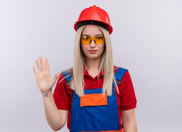 Довольная молодая блондинка инженер-строитель девушка в униформе в защитных очках, жестикулирующая на изолированном белом пространстве