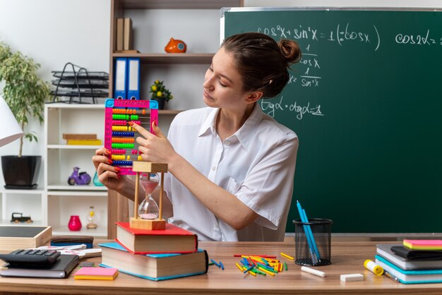 Довольная молодая блондинка учительница математики сидит за столом со школьными инструментами, держа, глядя и указывая пальцем на счеты в классе